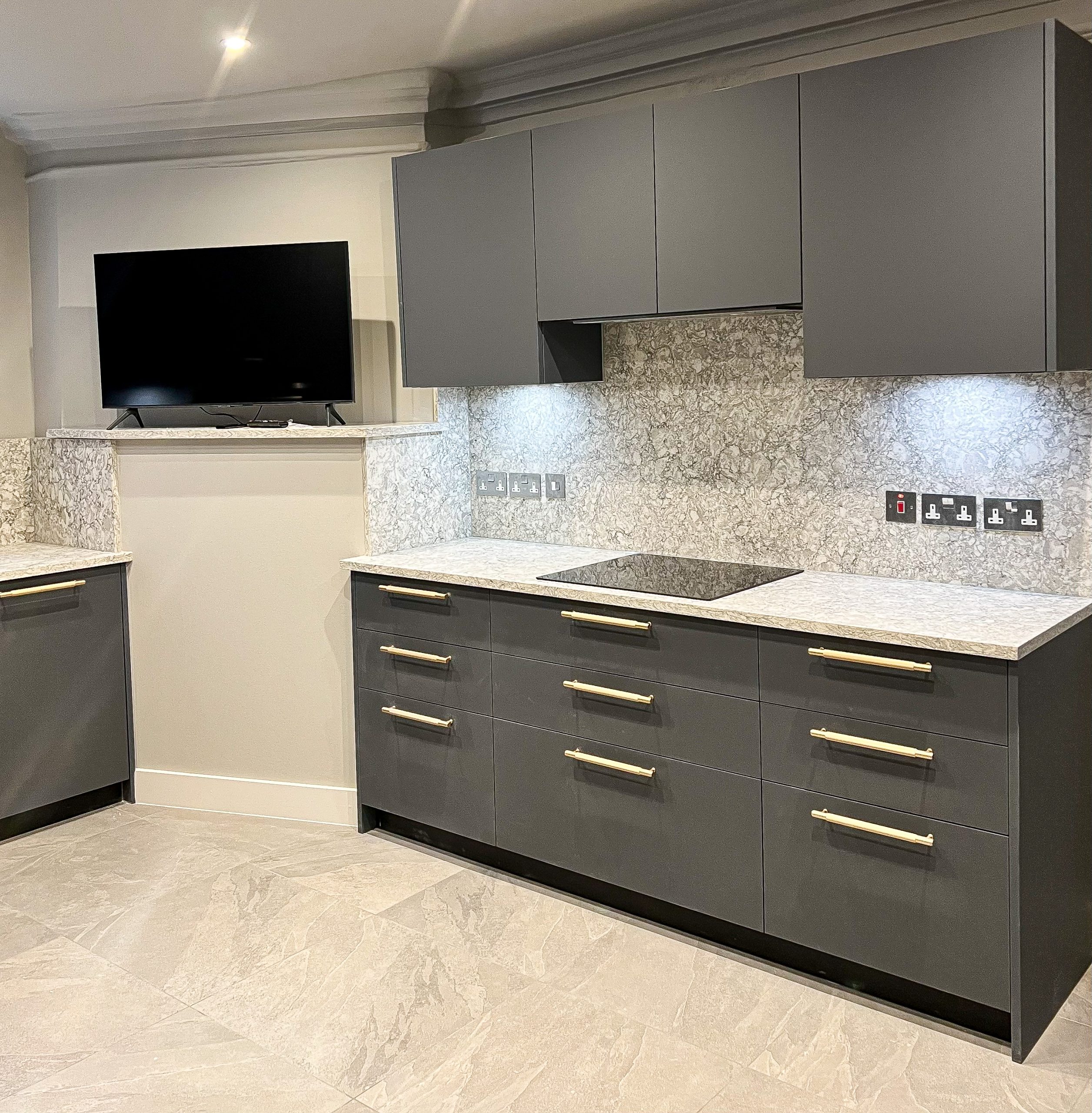 A modern kitchen with gray cabinetry, marble countertops, and a mounted television.