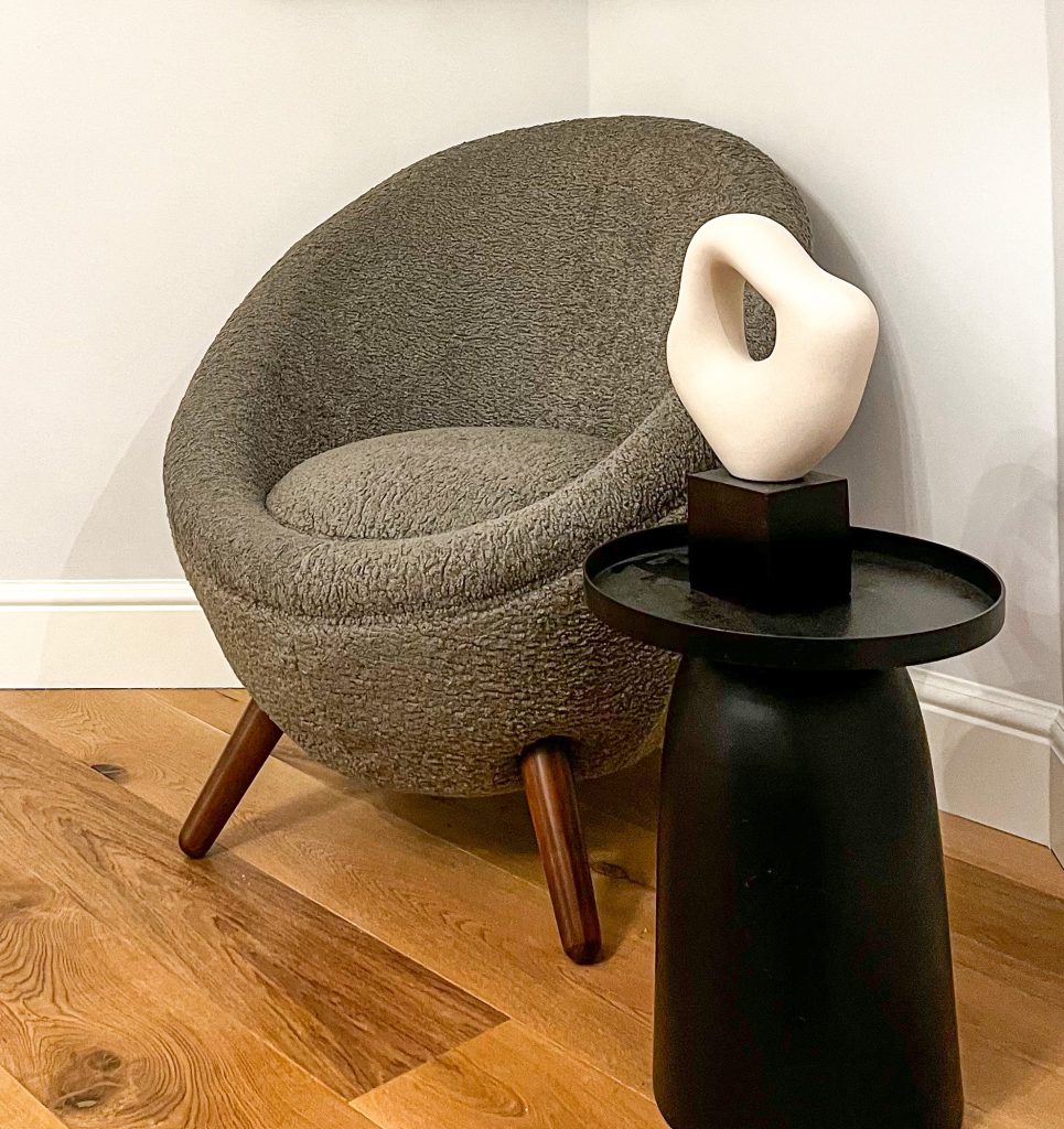 A modern textured round chair with angled wooden legs next to a black side table displaying an abstract white sculpture.